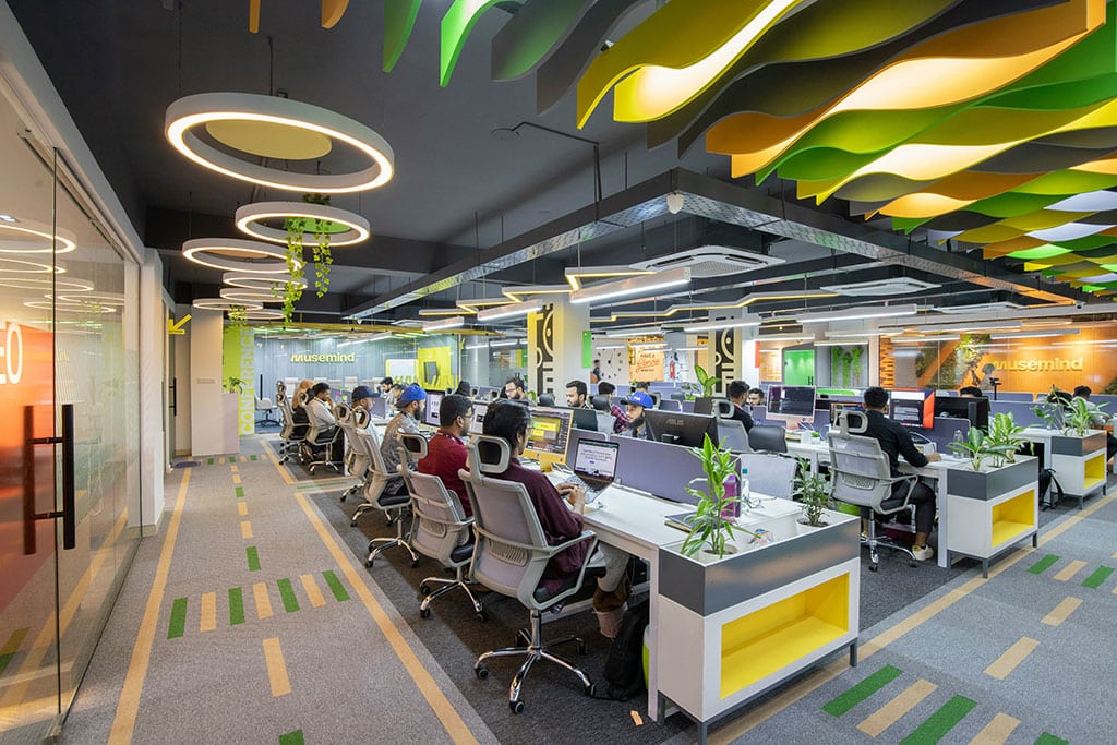 Image of a modern open-plan office with employees working at computer desks, featuring bright lighting, colourful ceiling designs, and indoor plants.