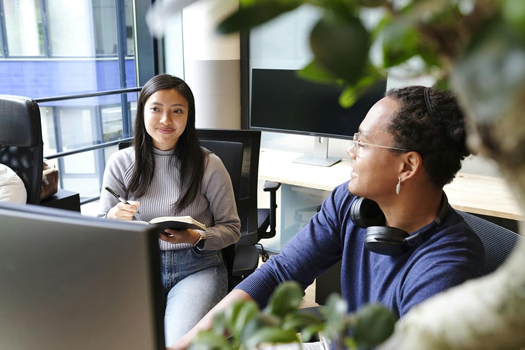 Image of two colleagues sitting at desks in a modern office, smiling and talking while one takes notes in a notebook.