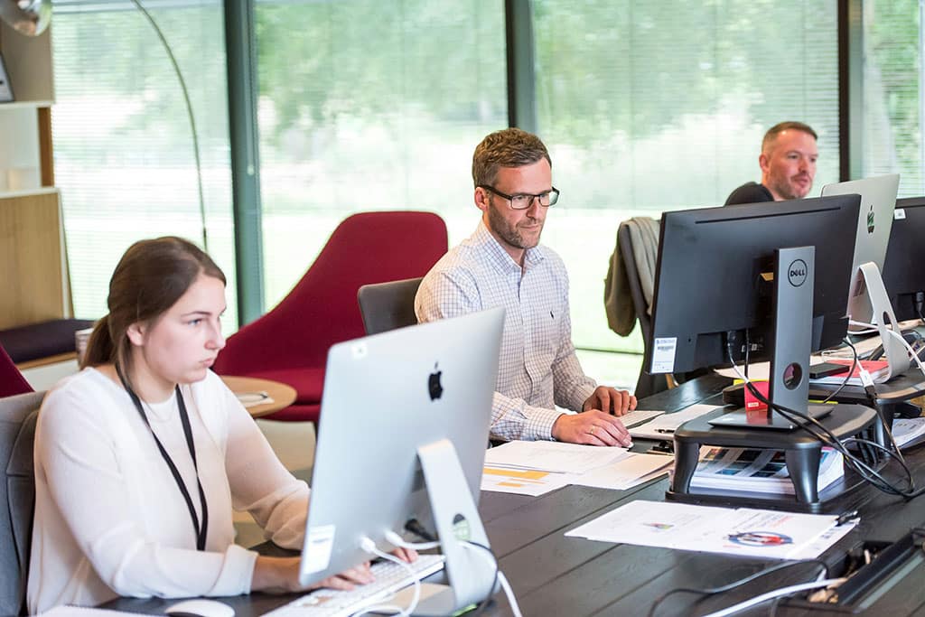 Image of three people working at computers in a modern office with large windows and natural light.