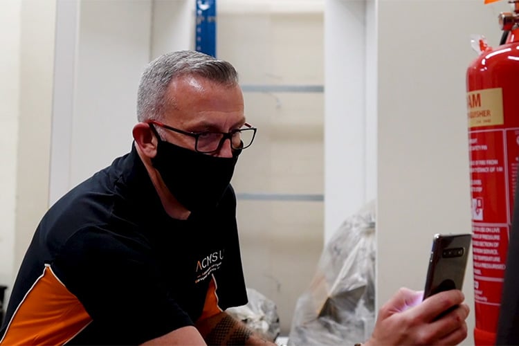 Image of a masked technician examining a fire extinguisher while referencing information on a smartphone in an indoor workspace.