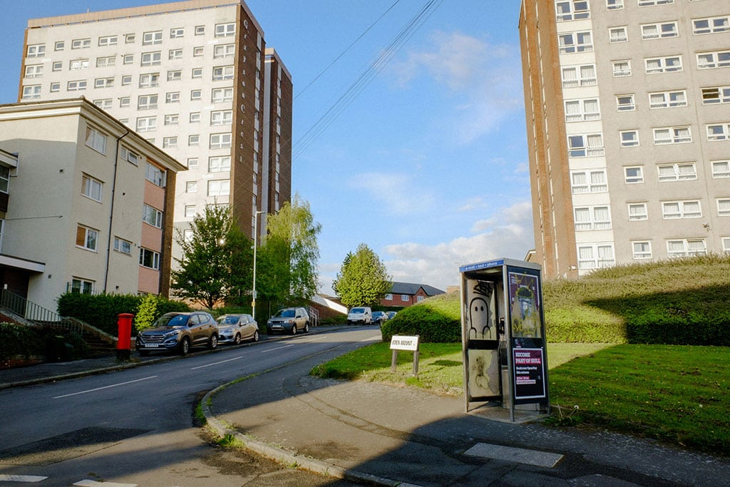 Image of a residential street with high-rise apartment buildings, parked cars, grassy areas, and a roadside phone booth under a clear blue sky.