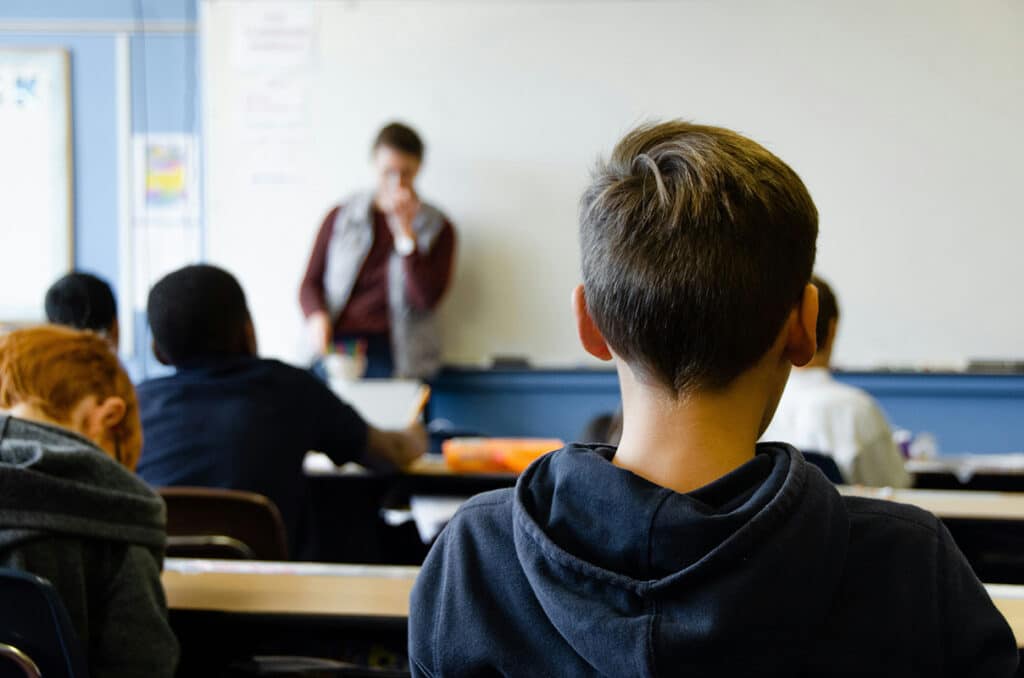 Image of a school classroom with students looking to the front and a teacher standing next to a white board, representing the idea of fire safety compliance in schools
