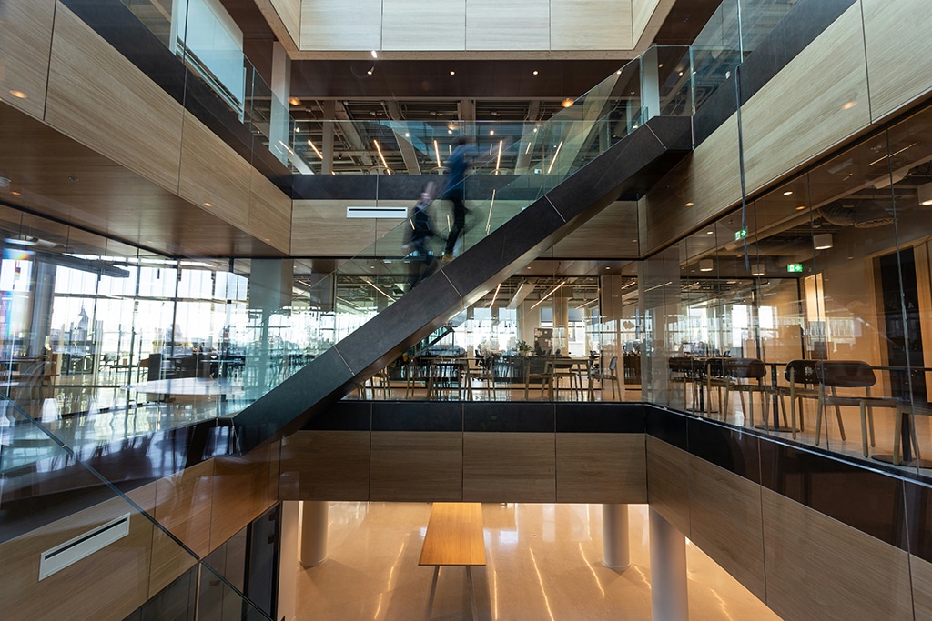 Image of modern office atrium with glass walls and wooden panels, person walking up central staircase above open seating area.