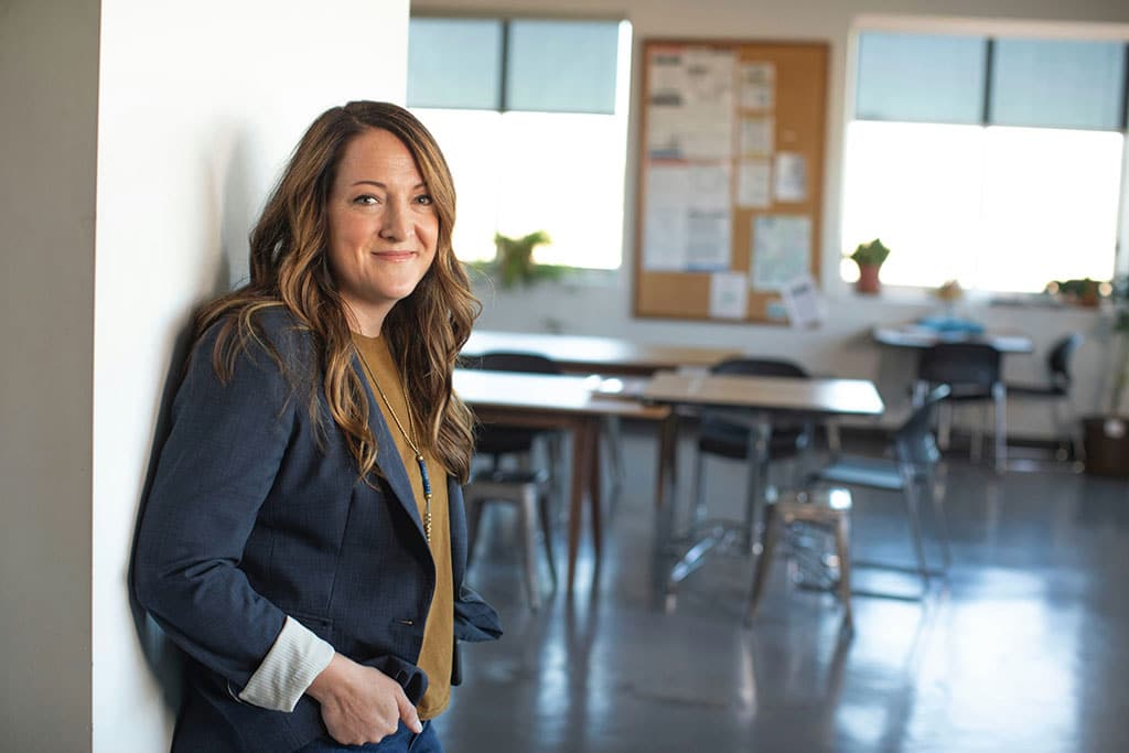 Image of a female school teacher leaning against a wall with classroom desks in the background and representative of the topic of School Condition Allocation.